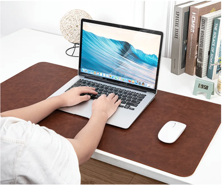 Person typing on a laptop placed on a NOIRDEN brown leather desk mat with mouse and books in background