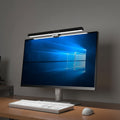 NOIRDEN Signature Monitor Light mounted on a white desktop monitor, casting warm white light on a wood desk with keyboard and mouse