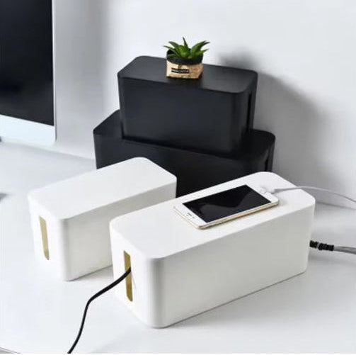 Two white rectangular cable management boxes with gold-colored interior slits, shown on a white desk with a smartphone resting on the front one. One box is open with a black cable plugged in, and the other is closed.