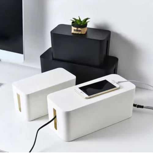 Two white rectangular cable management boxes with gold-colored interior slits, shown on a white desk with a smartphone resting on the front one. One box is open with a black cable plugged in, and the other is closed.